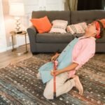 A man humorously poses with a broom while cleaning in a cozy living room.