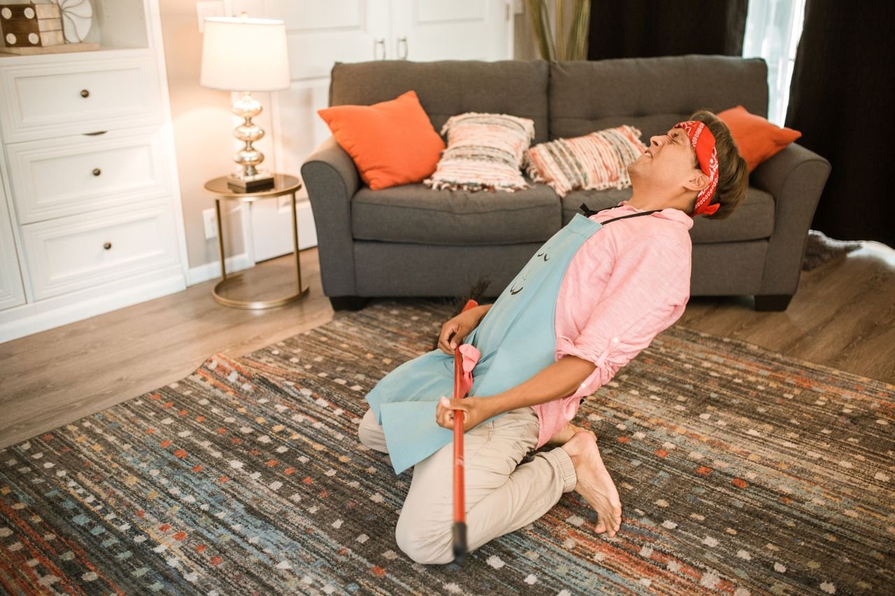 A man humorously poses with a broom while cleaning in a cozy living room.